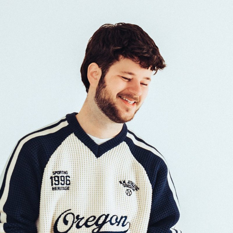 APRD, DJ y productor de Utrecht, sonriente con un suéter universitario de Oregon color crema sobre un fondo claro