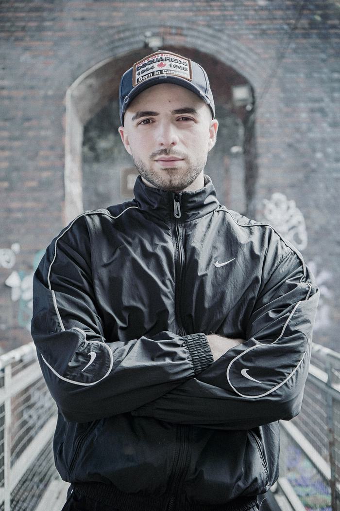 Jordan George, Hard-House-DJ aus Birmingham, mit Cap und schwarzer Trainingsjacke unter einem Backsteinbogen