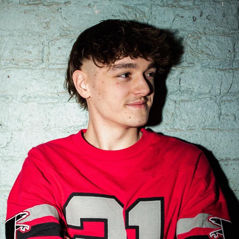 Leo Sanderson, House- und Rave-DJ aus Manchester, lächelnd im roten Trikot Nr. 21 vor heller Backsteinwand