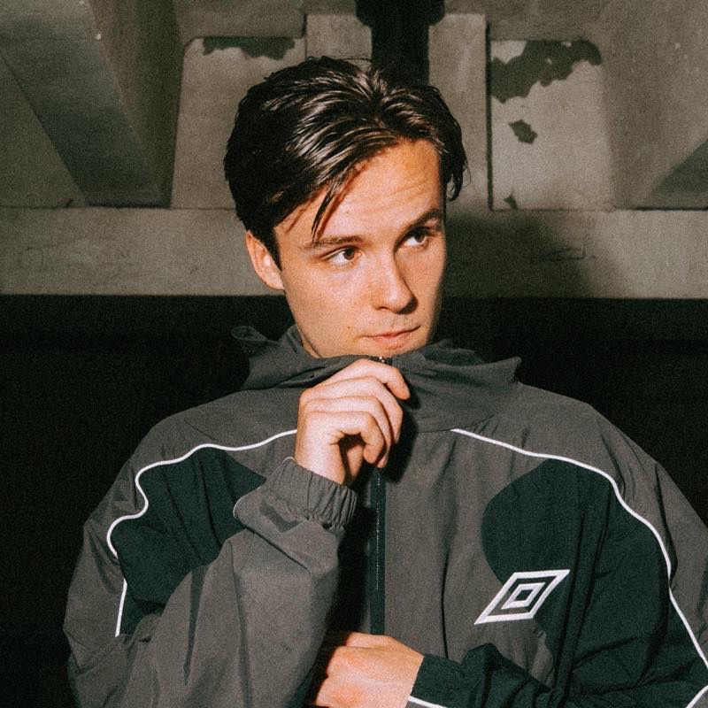 LUMINE, productor y DJ neerlandés, con chaqueta deportiva Umbro gris y negra ante una pared de cemento